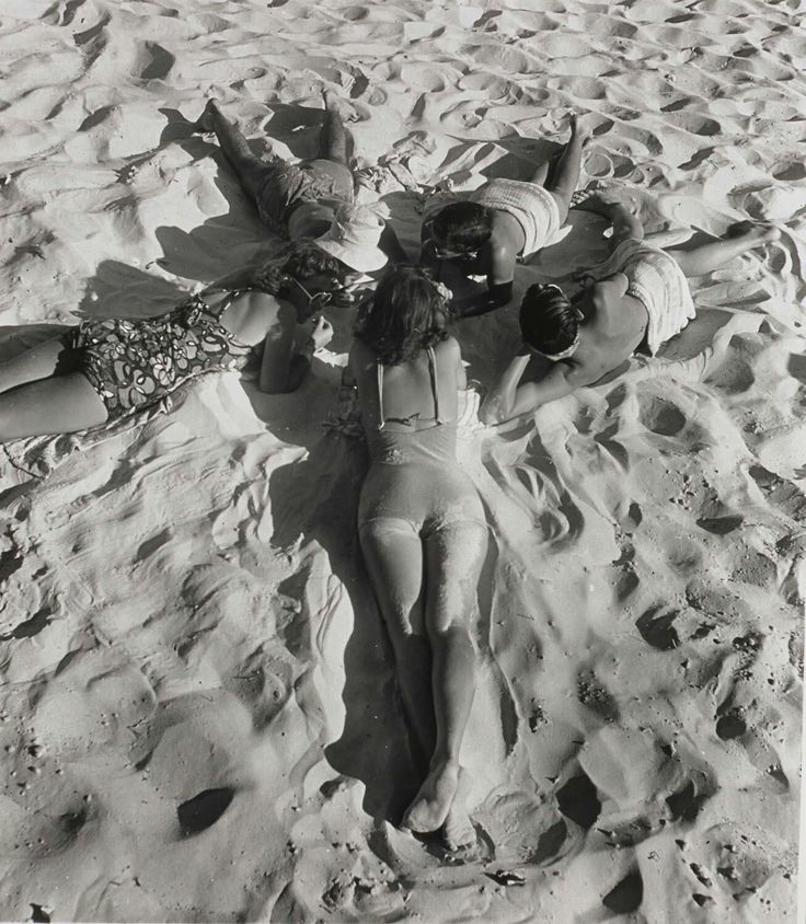 Five women in swimsuits lie in a circle on rippled beach sand, relaxing and chatting in a sunny black-and-white scene.