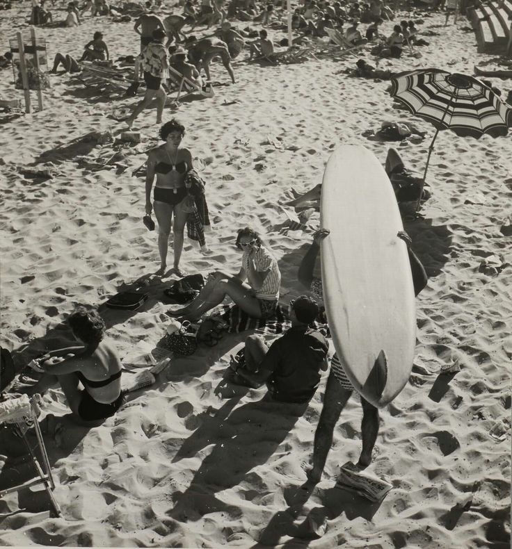 Crowded black-and-white beach scene with a person carrying a surfboard, sunbathers on sand, and a striped umbrella.