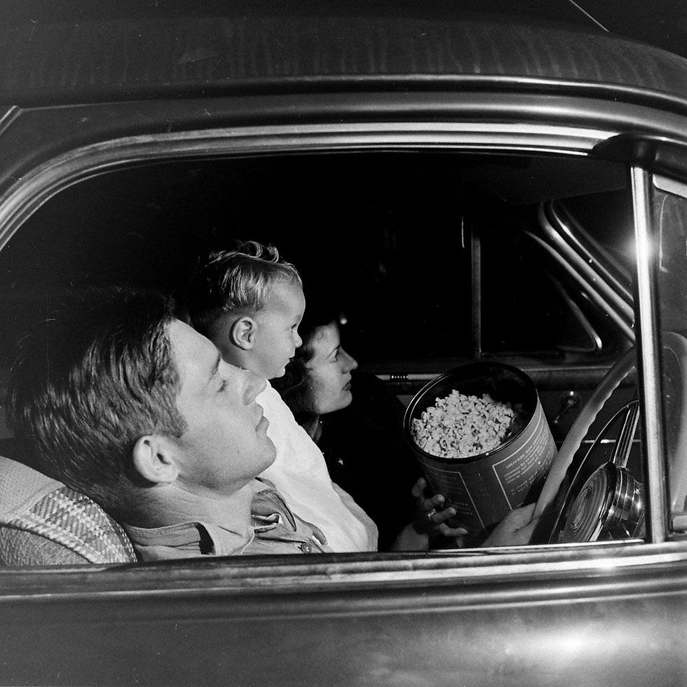 Black-and-white family in a car at drive in movie theatre, child on mother’s lap and holds a tub of popcorn.