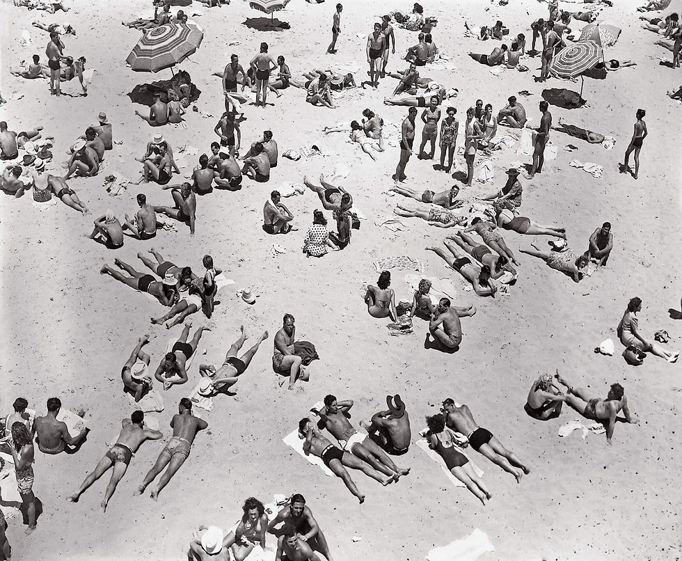 Crowded black-and-white beach scene with dozens of sunbathers lounging on towels, chatting, and standing under striped umbrellas.