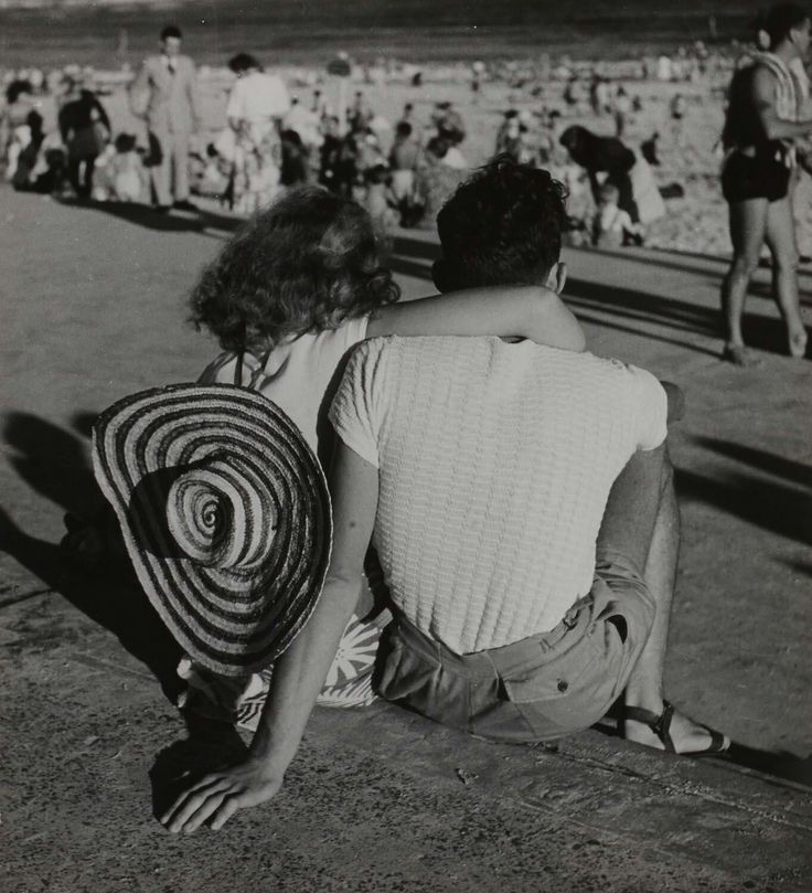 Black-and-white couple sits on a seaside promenade, woman in a striped hat hugging a man, with beachgoers blurred behind.