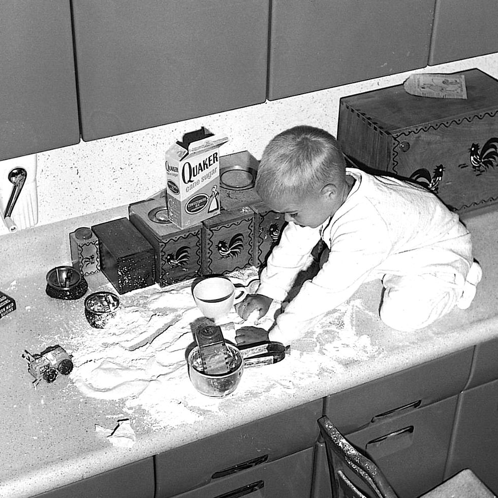 Toddler kneels on kitchen counter playing in spilled sugar beside a  sugar, flour and measuring cups.
