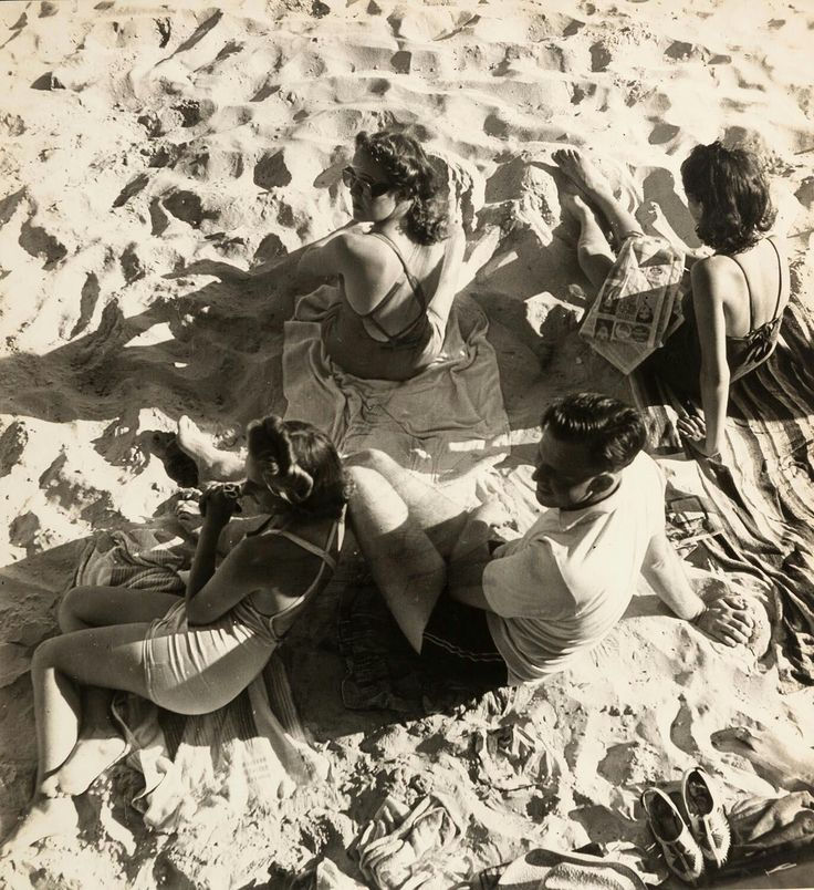 Black-and-white overhead view of four people relaxing on a sandy beach, sitting on towels and chatting; a magazine and shoes nearby.