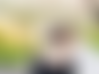 Von hinten aufgenommene Fotografie einer jüngeren Frau, die einen Arm um eine im Rollstuhl sitzende, ältere Person mit grauem Haar legt, während beide in einen sonnigen Park mit blühenden Bäumen blicken.