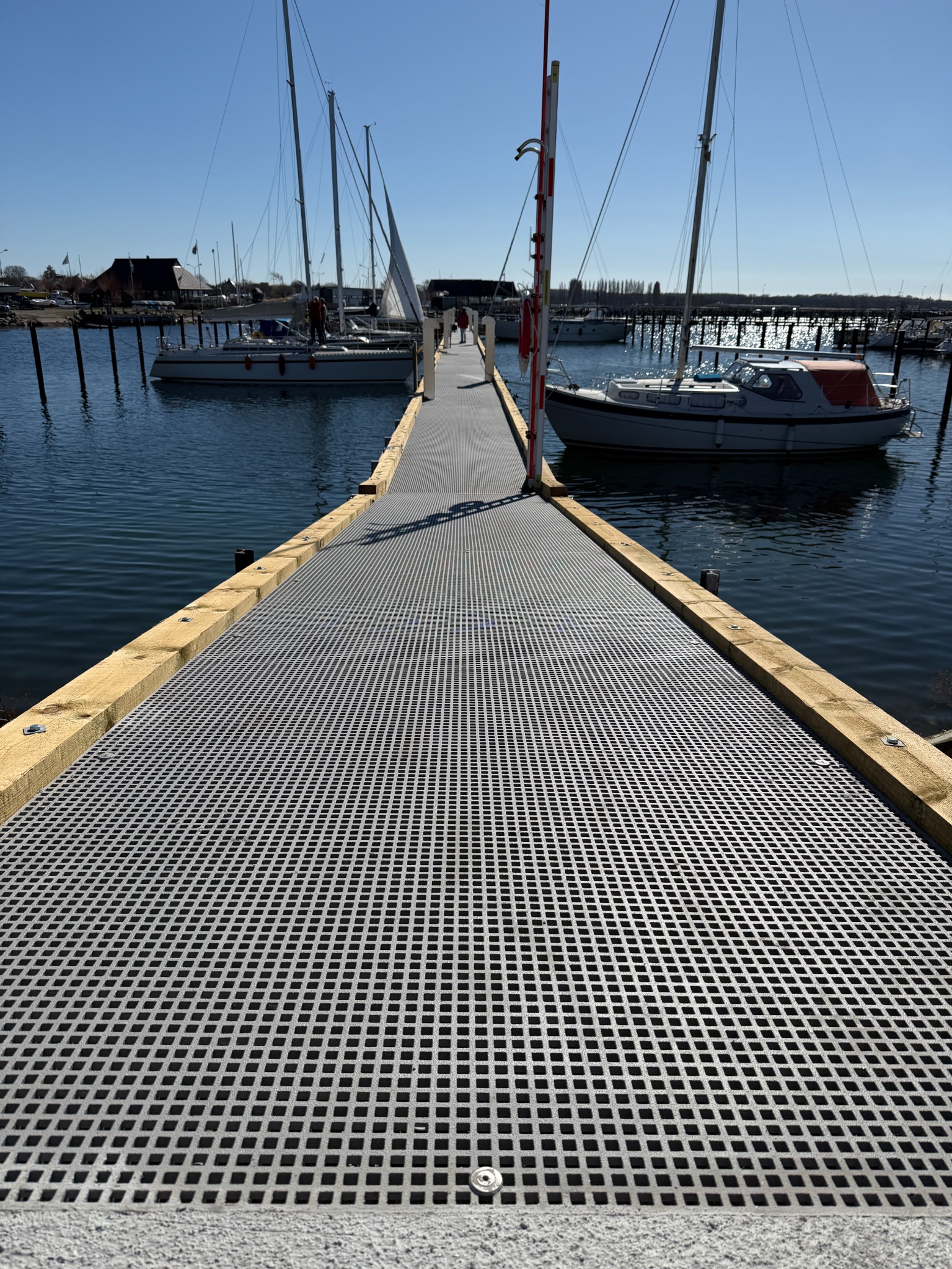 Pier with grey GRP grating between moored sailboats in a marina