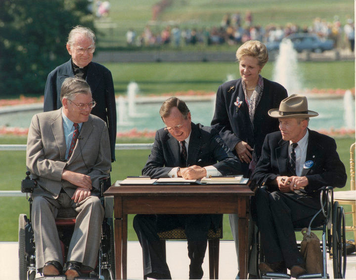 President George H. W. Bush signing the ADA at desk outdoors with four individuals on either side, two of whom are seated in wheelchairs.