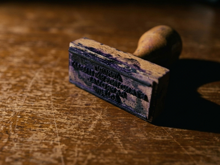 A worn rubber stamp lying face up on a scratched wooden desk, its ink-stained face too blurred to read