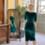 Model in a green velvet dress stands before a mirror in an elegant room with a plant and wooden furniture, reflecting a serene mood.