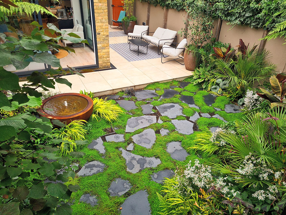 Lush garden with stone pathway, green plants, and a round water fountain. Cozy patio with white chairs in the background. Calm atmosphere.