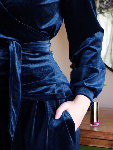 Close up of a shimmering blue velvet top featuring tie waist with model's hand casually slipped into the trouser pocket, showing opulent elegance