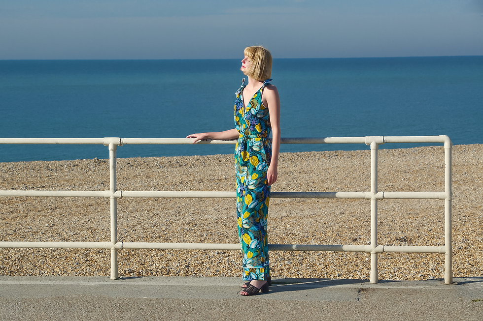 A person in a colorful jumpsuit stands beside a railing, gazing at the sea on a sunny day. Pebble beach and blue ocean in the background.
