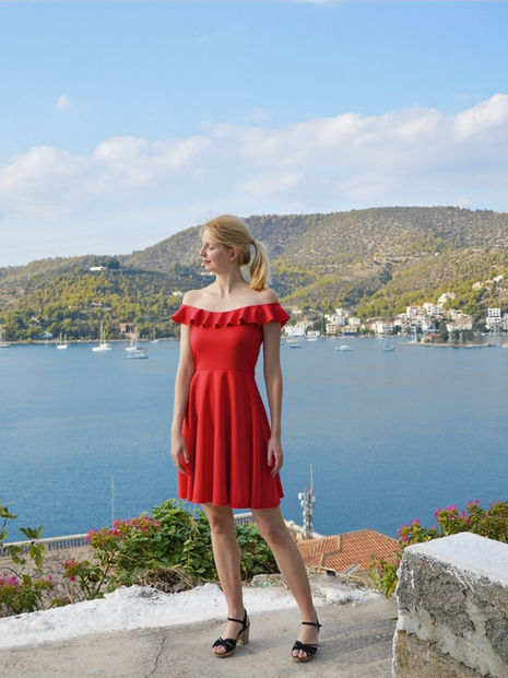 Woman standing on a sunlit terrace overlooking Poros bay, wearing the Ginger off-shoulder skater dress in red, paired with black wedge sandals