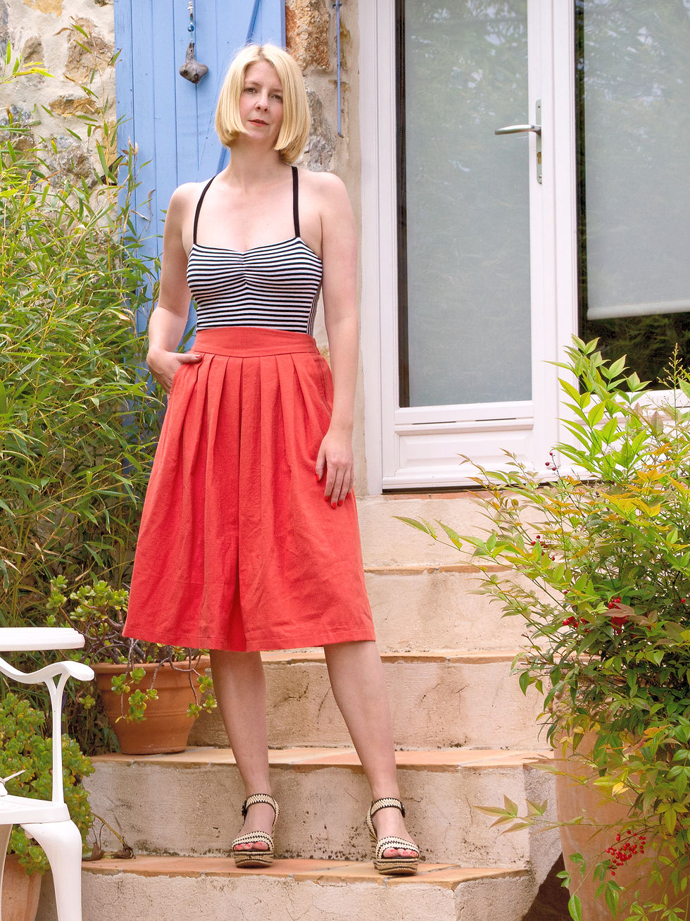 Woman with blonde bob stands on stone steps, wearing striped top and coral orange skirt. Blue shutters and greenery in background, relaxed mood.