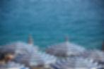 Blue and white striped beach umbrellas on Cote d'Azur seafront partially cover people on a pebbled shore. Calm turquoise sea in the background.