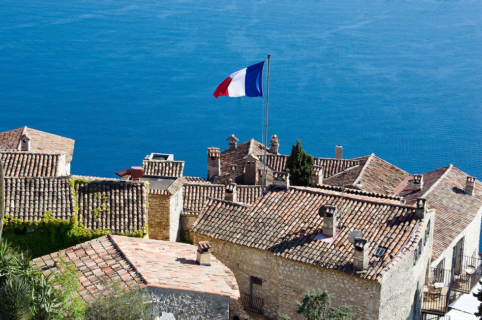 French flag waving over terracotta rooftops of a coastal village Eze with bright blue sea in the background.