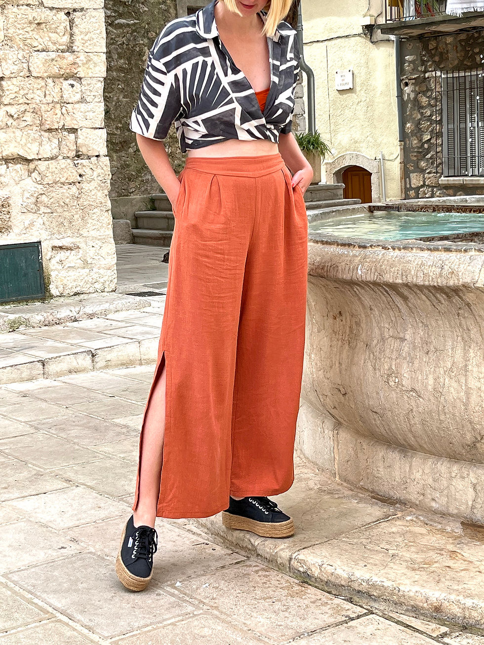 Model in black-and-white shirt and orange wide leg trousers stands by a stone fountain in a sleepy hilltop village. Serene holiday mood and styling