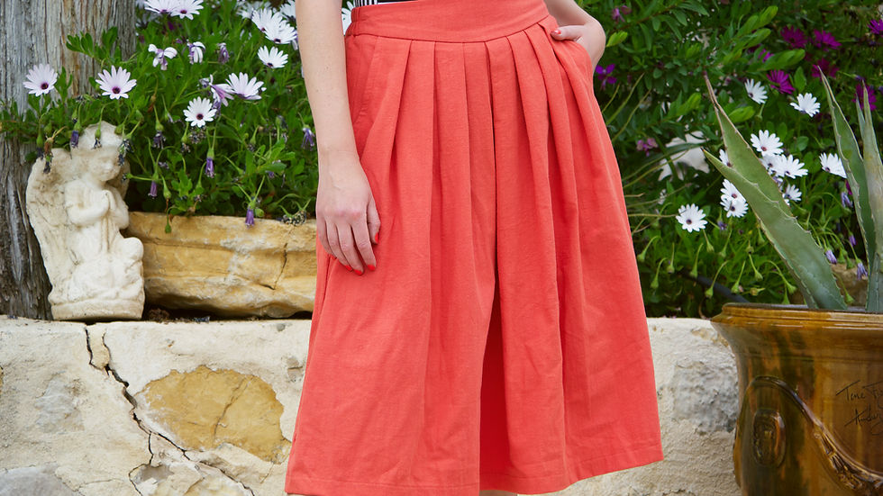 Woman in a striped top and orange skirt stands by flowers and a stone statue, hands in pockets. Rustic stone backdrop, lush greenery.
