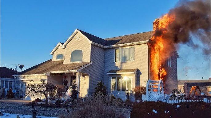 Two-story house with fire engulfing the chimney. Bright blue sky, sunlight on windows. Nearby bushes and snow. No visible people.