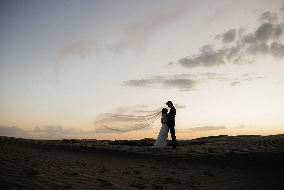 A romantic silhouette of a couple standing on sand dunes at sunset. The bride's veil flows dramatically in the wind, creating a dreamy and ethereal atmosphere.