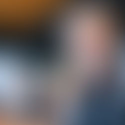 Ein selbstsicher wirkender älterer Mann mit grauem Bart und Brille sitzt in einem modernen Meetingraum und blickt ruhig nach vorn. Seine entspannte, konzentrierte Haltung vermittelt deutlich Selbstbewusstsein als Führungskraft, während im unscharfen Hintergrund weitere Teammitglieder sitzen.