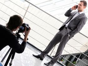 Fotograf fotografiert einen Geschäftsmann im grauen Anzug während eines professionellen Business-Shootings in einem hellen Gebäude mit Glasdach und Metallgeländer.