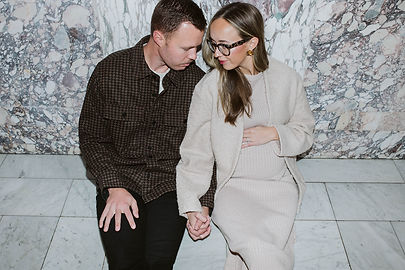 A couple holding hand and sitting on a tile floor with their backs against a granite wall. The woman is pregnant.