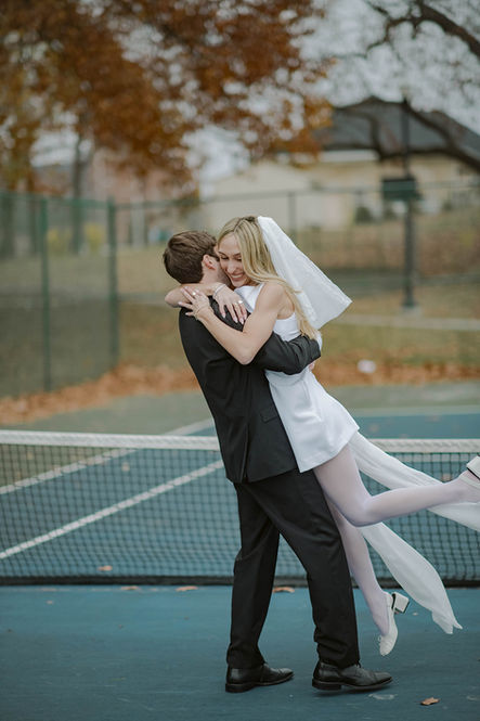 An outdoor portrait of a man lifting a woman into the air for a playful embrace on an outdoor court, with autumn trees visible in the background.