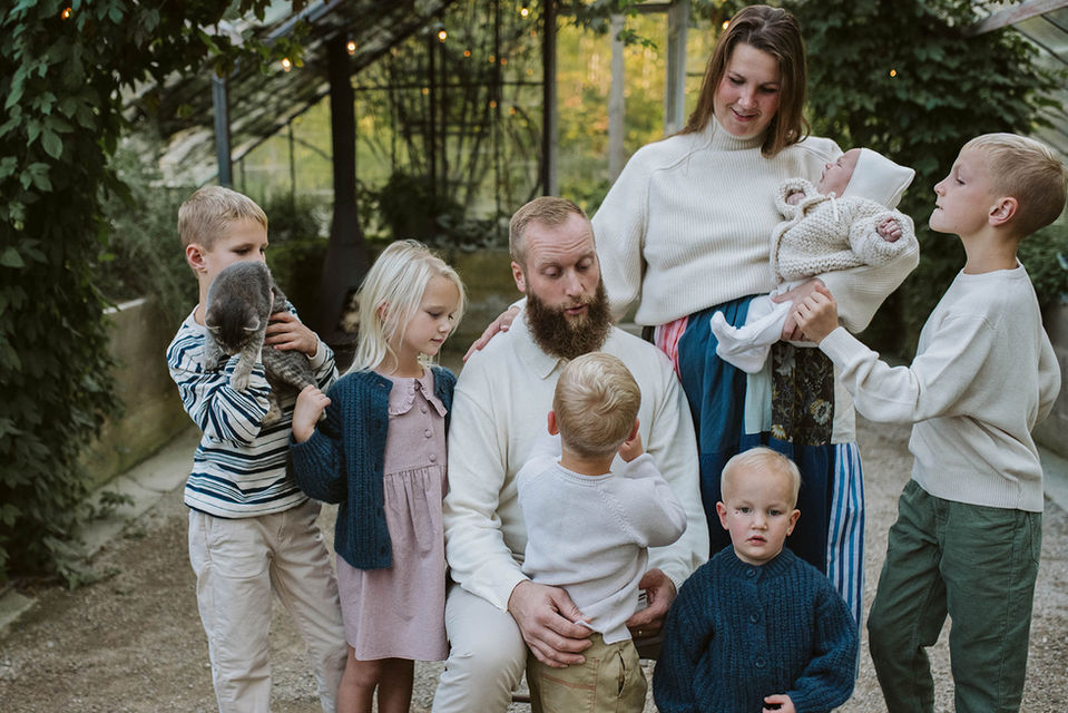 A close-up group portrait of a father holding a baby, surrounded by his other four children as they smile together in front of the greenhouse.