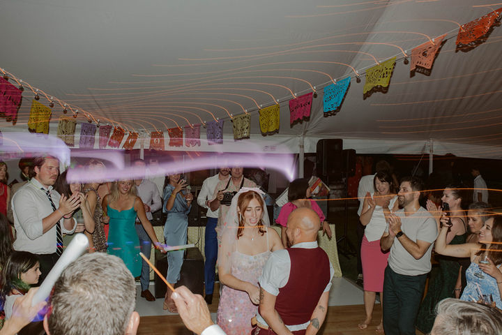 A lively wedding reception scene showing guests dancing under a tent ceiling decorated with colorful, festive bunting banners.