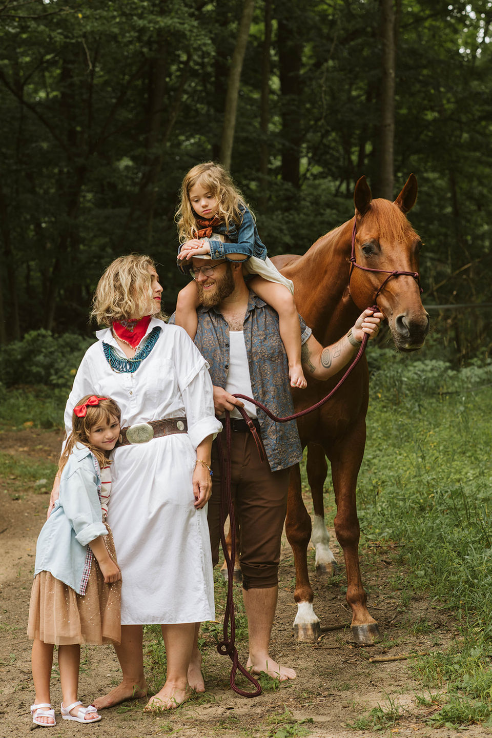 A woman and three young children standing together on a dirt path next to a brown horse in a rustic, wooded setting.