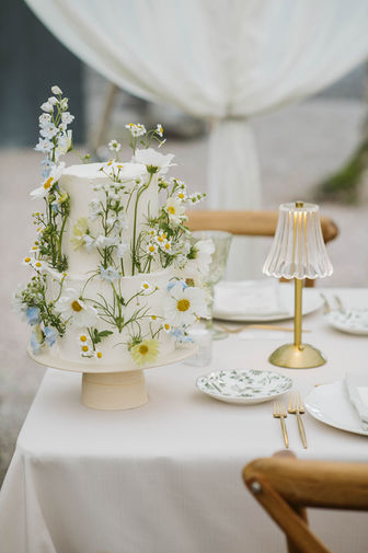 A close-up of an elegant outdoor wedding reception table setting, featuring a white floral centerpiece, burning taper candles, and minimalist dinnerware.
