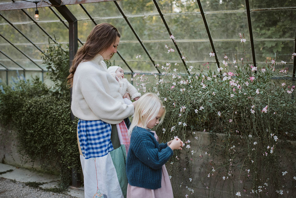 A mother holding a baby while standing next to her young daughter outside a greenhouse wall lined with pink flowers.