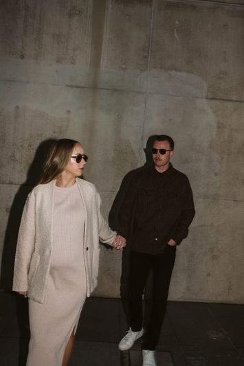 A man and woman walking together at dusk or in a shadowed urban area, with soft architectural lighting in the background.