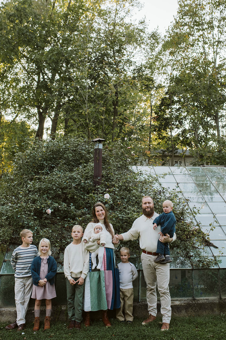 A full family portrait of a mother, father, and their five children standing together outside in front of a large, glass greenhouse structure surrounded by green trees.