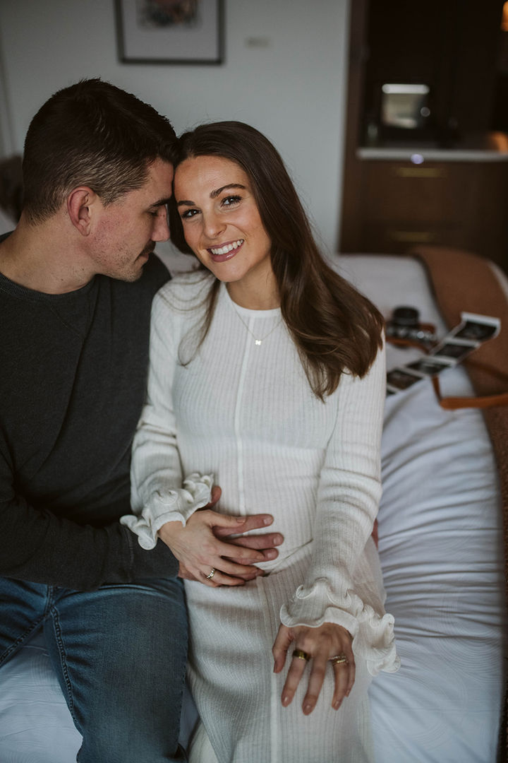 A candid moment of the couple sitting on a bed together, smiling as they look down at ultrasound photos of their baby.