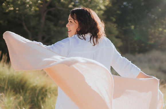 Smiling woman, who is the owner, in white shirt with flowing shawl behind her.  She is on the beach with dune grass and trees in the background.