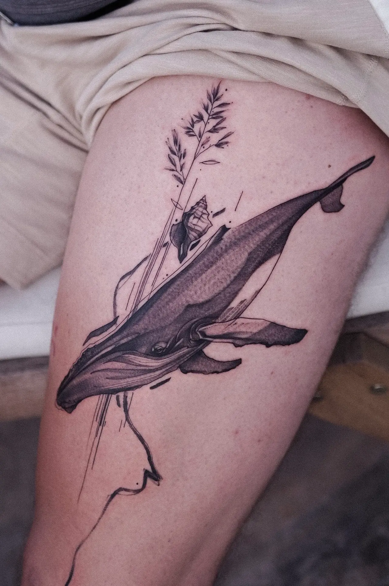 Schwarzgraues Buckelwal-Tattoo am Oberschenkel, frisch — Fine-Line-Akzente, kleine Muschel und Gräser.