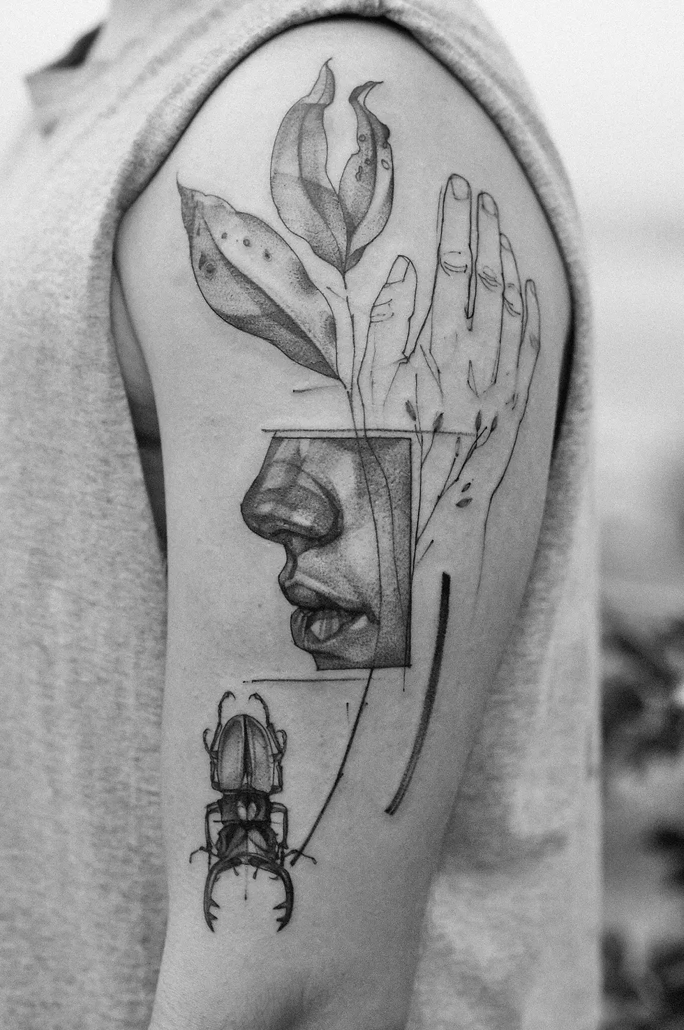 Black-and-white tattoo on upper arm with leaves, a hand, a profile face, and a beetle in close-up.