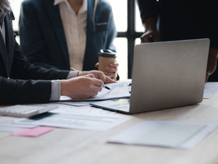 Twee mensen werken aan een tafel met laptops en papieren. Een persoon wijst met een potlood op een document. Rustige kantooromgeving.