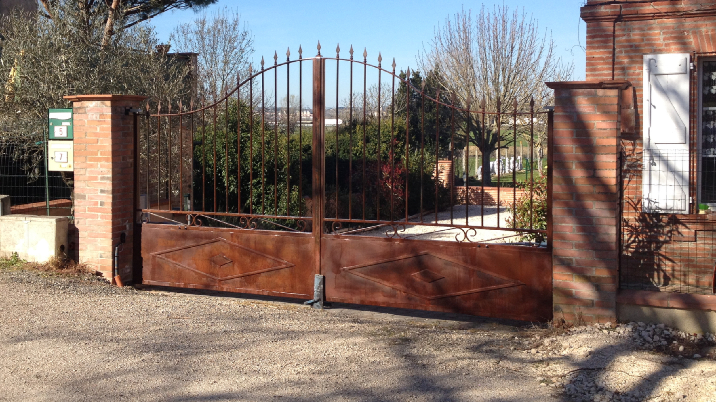 Une porte rouillée ouverte, un bâtiment en briques et la végétation dans le jardin.