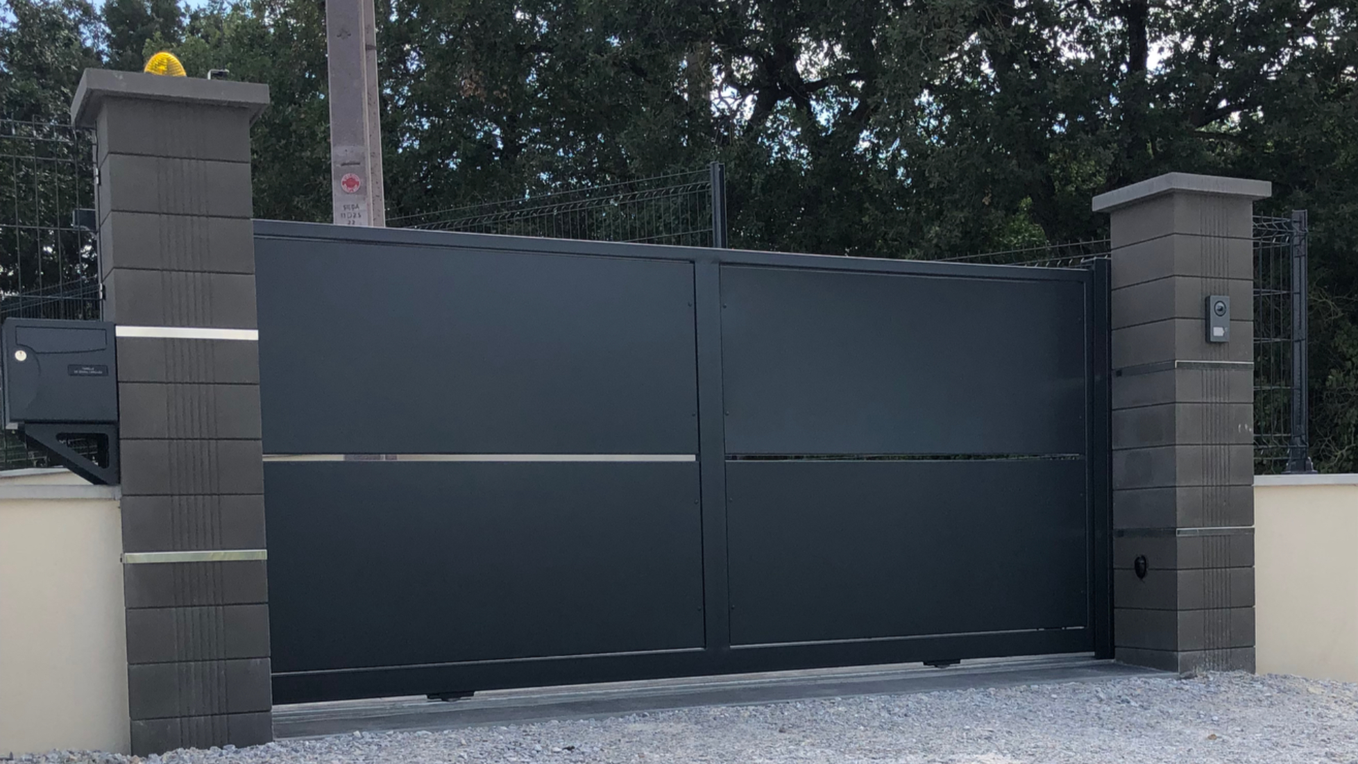 Un portail coulissant gris, avec des piliers, texte visible, et des arbres en arrière-plan.