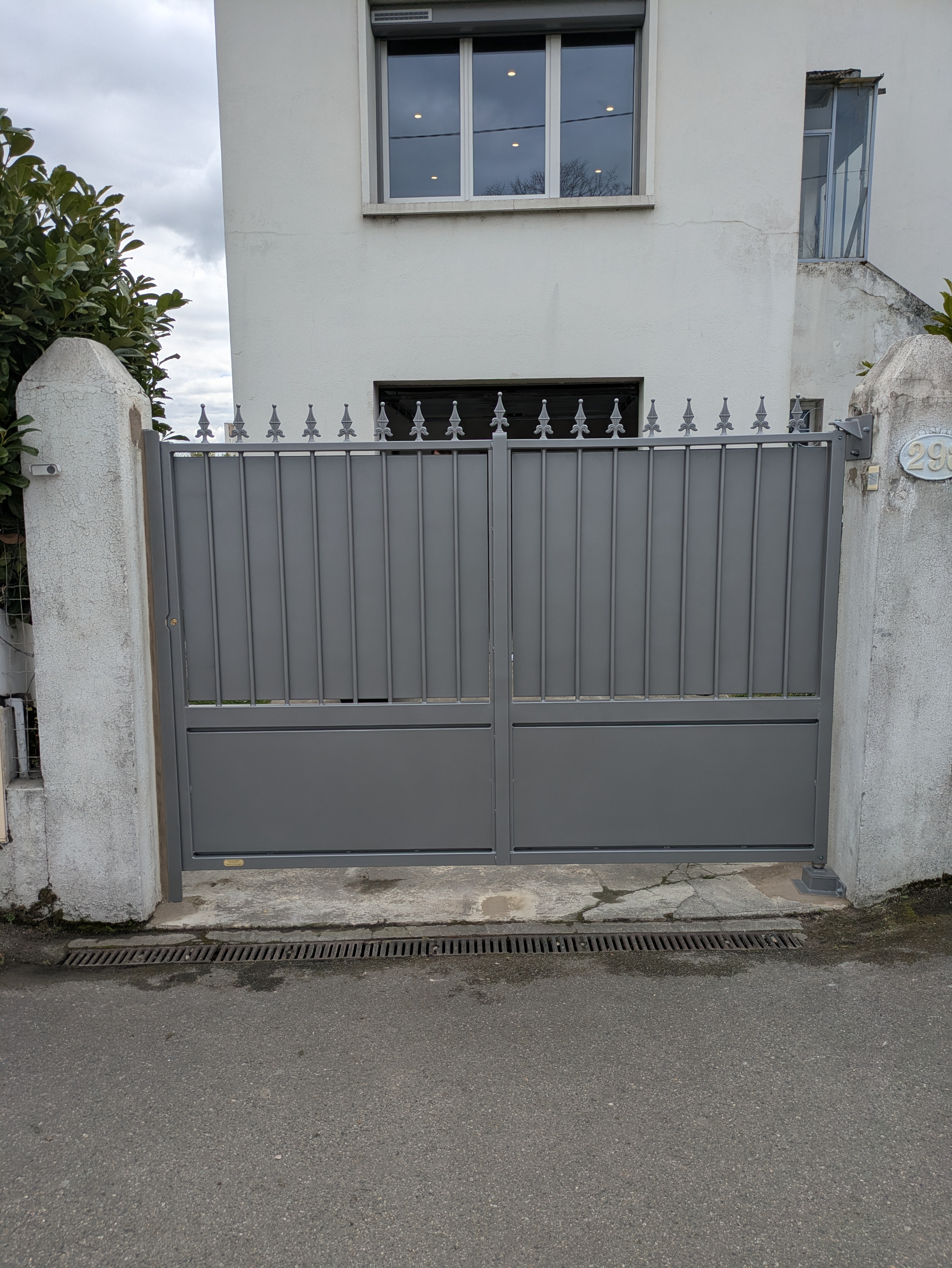 Portail gris fermé devant une maison blanche, avec des fenêtres et arbres.