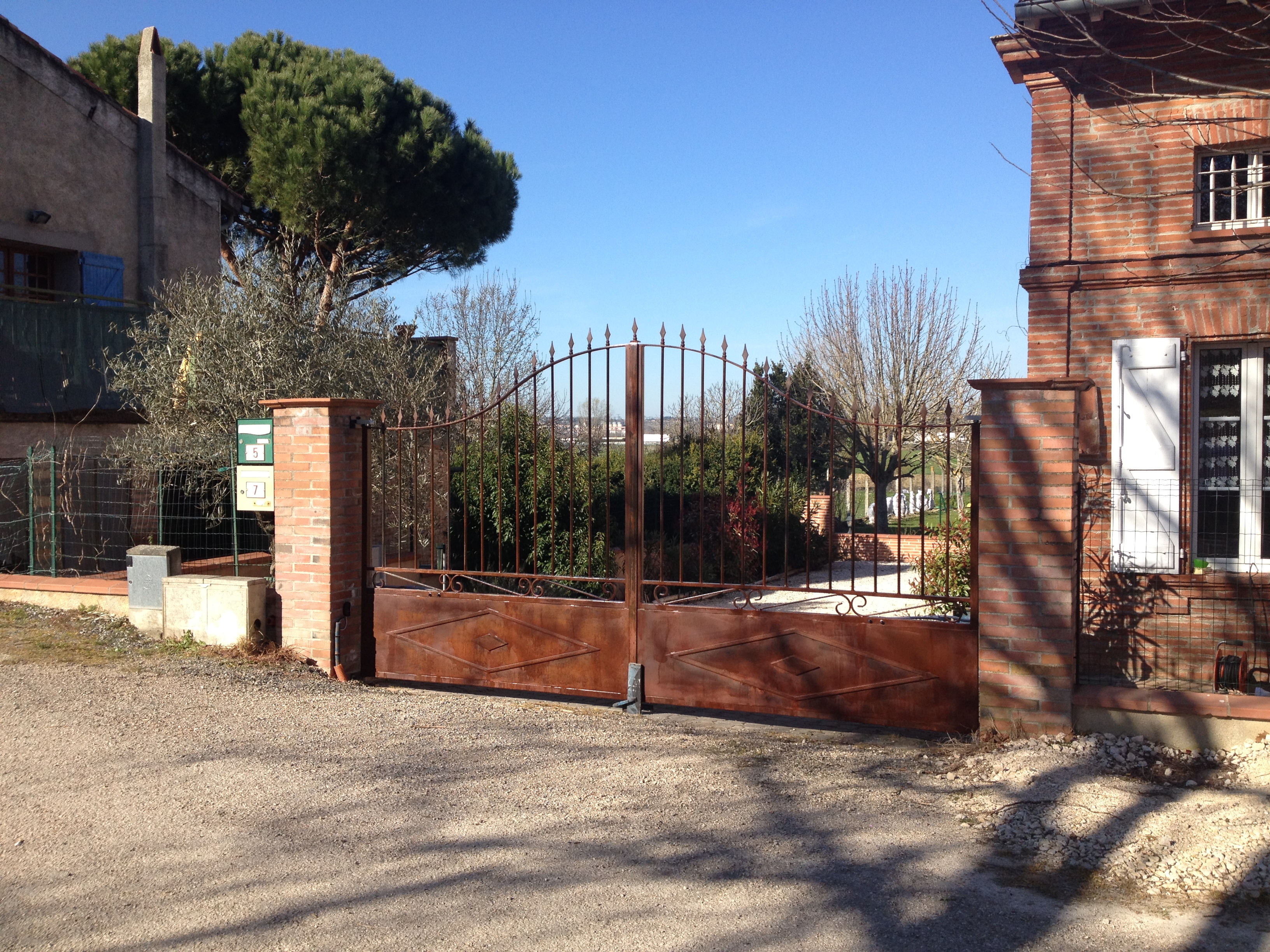 Une porte rouillée ouverte, un bâtiment en briques et la végétation dans le jardin.