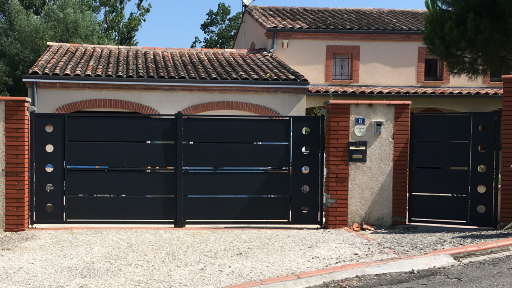 Maison avec portail noir. Mur en briques et arbres en arrière-plan, jour ensoleillé.