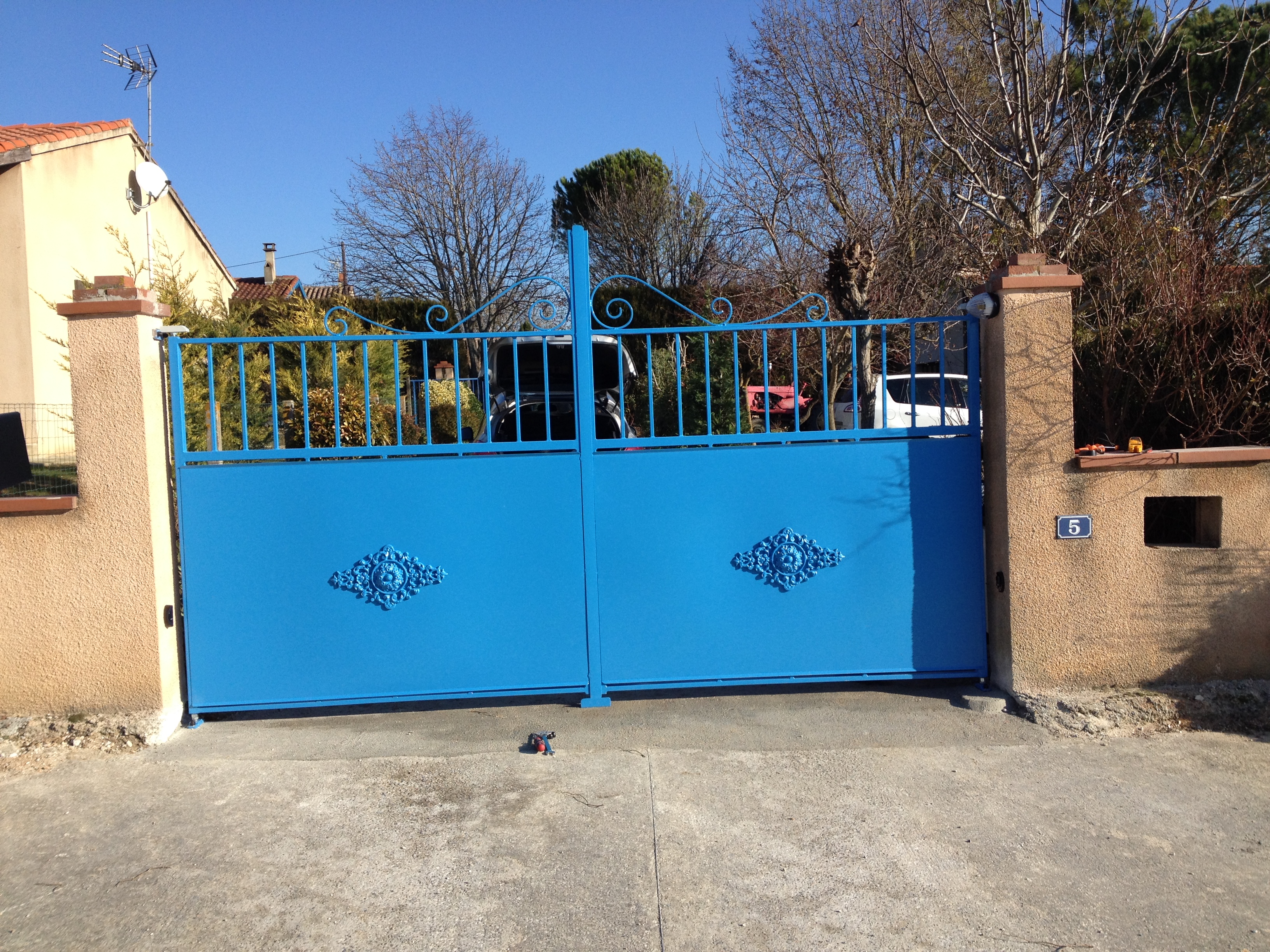 Portail bleu fermé devant une maison. Décorations et texte avec numéro.