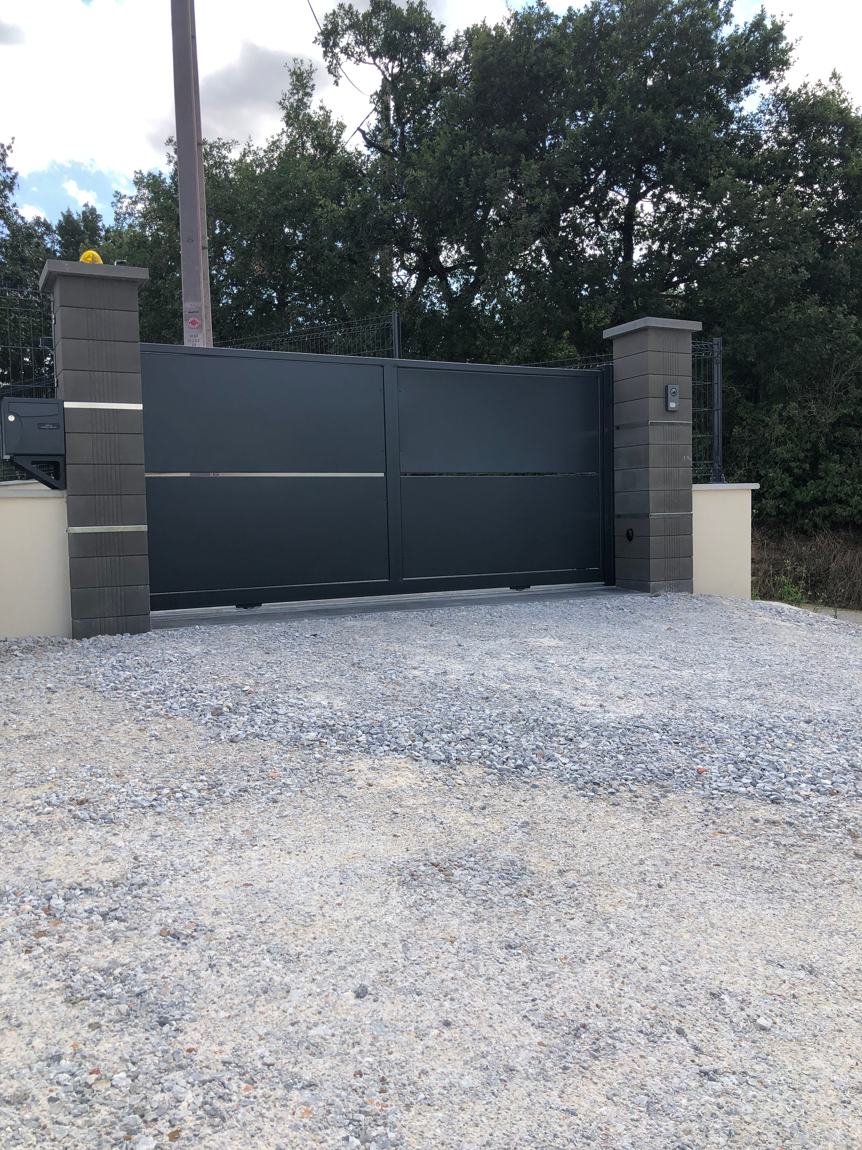 Un portail coulissant gris, avec des piliers, texte visible, et des arbres en arrière-plan.