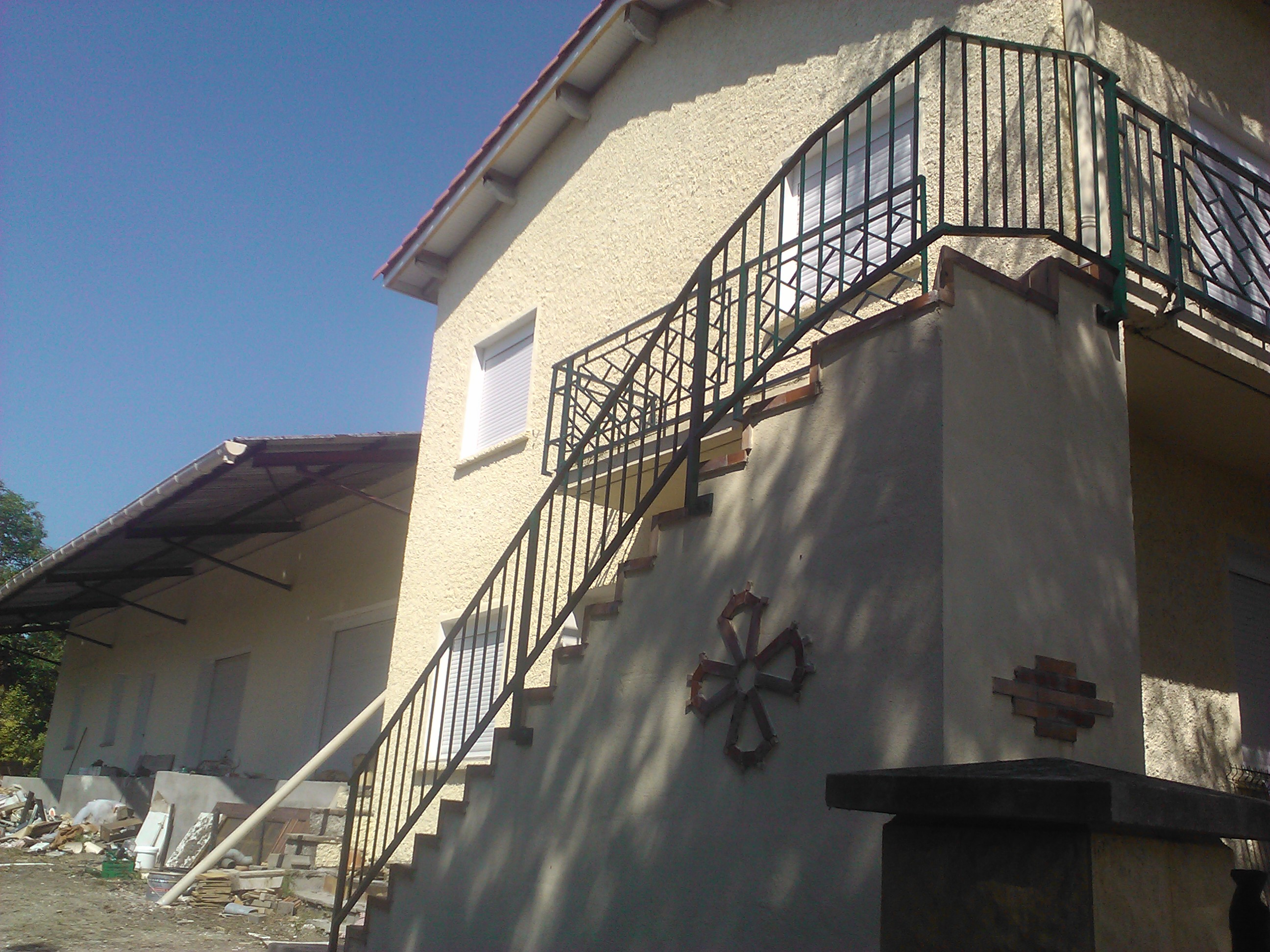 Escalier extérieur avec rampe en métal noir et une croix décorative sur le mur.