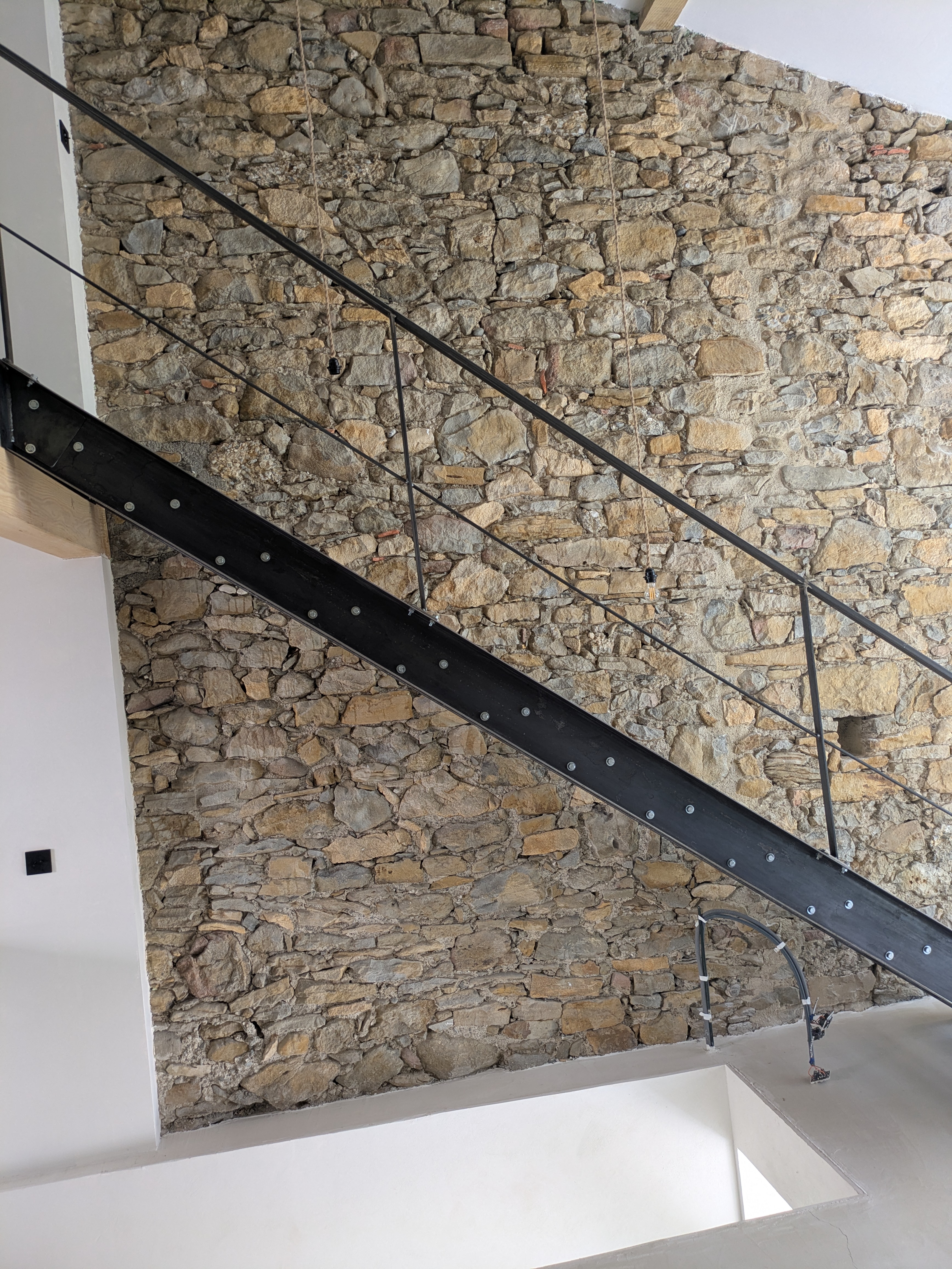 Escalier et rampe en métal noir devant un mur de pierres apparentes, Christo.