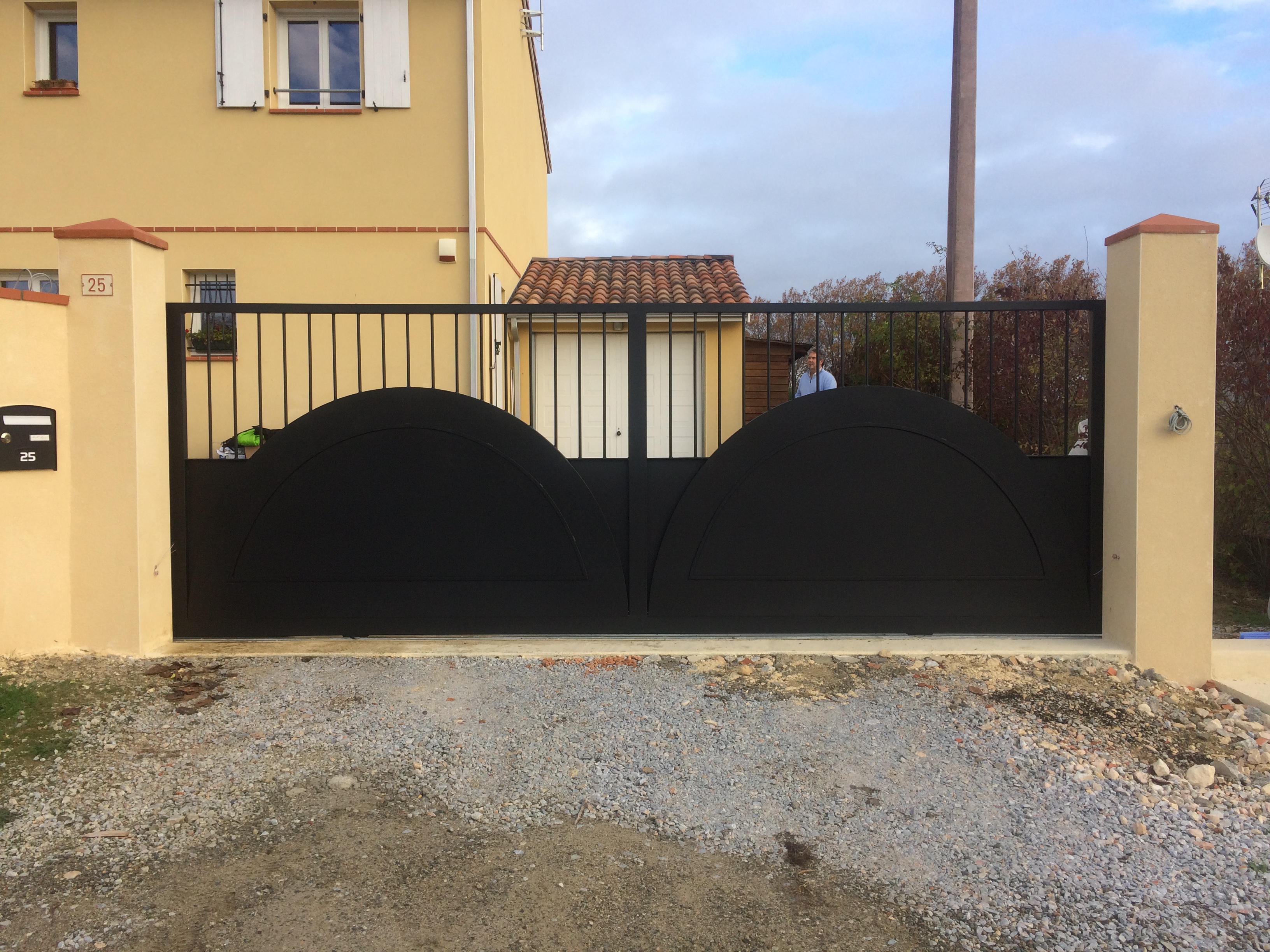 Portail noir incurvé devant une maison jaune avec des détails beiges.