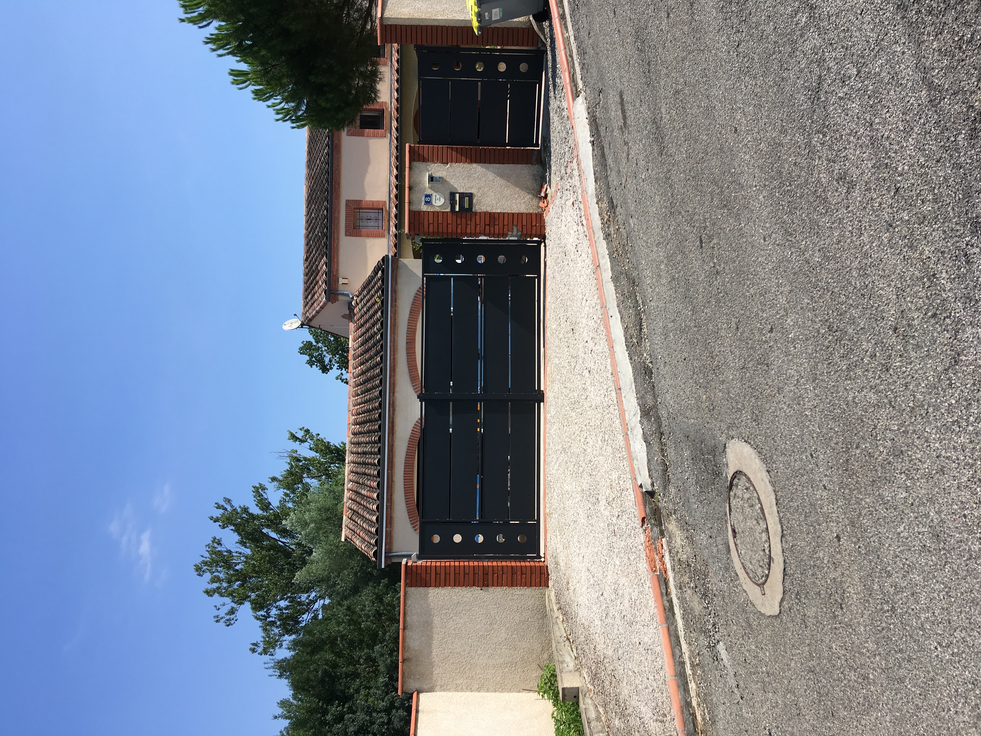 Maison avec portail noir. Mur en briques et arbres en arrière-plan, jour ensoleillé.